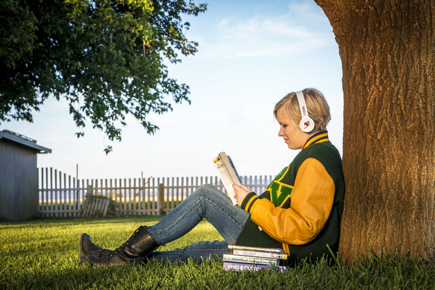 A high-school senior in a letterman jacket and headphones reads under a tree at golden hour.